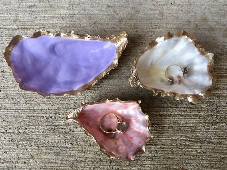 K&ouml;nnte beinhalten: Drei Ringteller aus Austernmuscheln, jede in einer anderen Farbe bemalt. Die erste ist lila, die zweite wei&szlig; und die dritte rosa. Jede Schale hat einen goldenen Rand und ist mit einem goldenen Maldesign verziert.