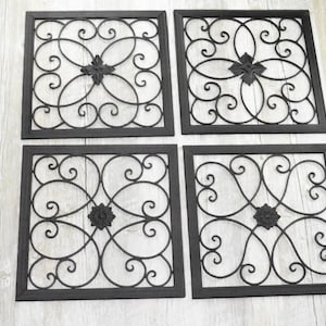 Può includere: Quattro pannelli quadrati decorativi da parete in metallo nero. Ogni pannello presenta un design decorativo e simmetrico con linee a spirale e un accento floreale centrale. I pannelli sono posizionati su uno sfondo chiaro con venature del legno.