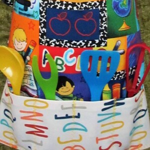 May include: A colorful patchwork apron with a pocket filled with yellow, green, and blue kitchen utensils. The apron features a variety of school-themed fabric squares with images of apples, globes, and children. The pocket has the alphabet printed on it.