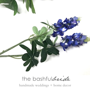 May include: Artificial bluebonnet stems with green leaves and the text "the bashful bride handmade weddings + home decor" on a white background. The bluebonnet flowers have a mix of blue and white petals.
