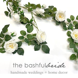 May include: White artificial rose garland with green leaves. The garland is arranged in a decorative pattern on a white background. The text "the bashful bride handmade weddings + home decor" is displayed below the garland.