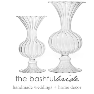 May include: Two clear glass vases with a ribbed design. The larger vase is taller and wider than the smaller vase. Both vases have a flared opening and a round base.