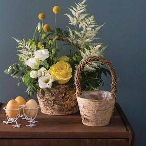 May include: Two birch bark baskets with wicker handles, one filled with white and yellow flowers, the other empty. The baskets are on a wooden table with three white egg holders and yellow eggs.