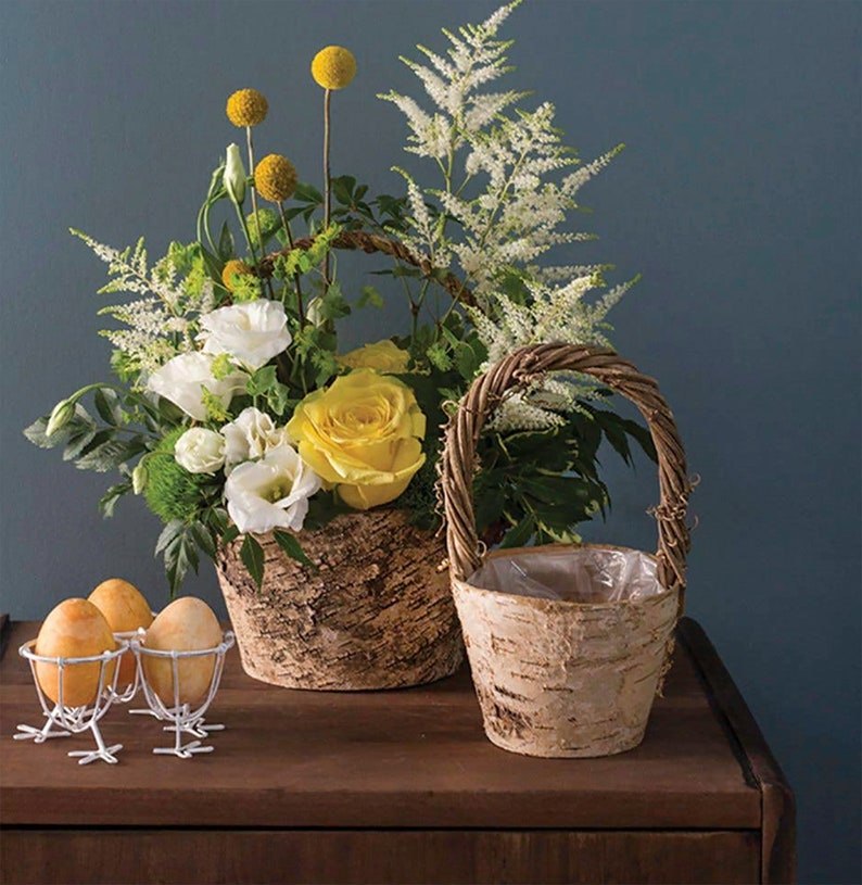 May include: Two birch bark baskets with wicker handles, one filled with white and yellow flowers, the other empty. The baskets are on a wooden table with three white egg holders and yellow eggs.