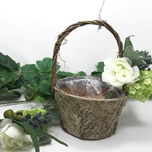 May include: A small, brown, bark-textured basket with a twig handle. The basket is filled with white flowers and green foliage.