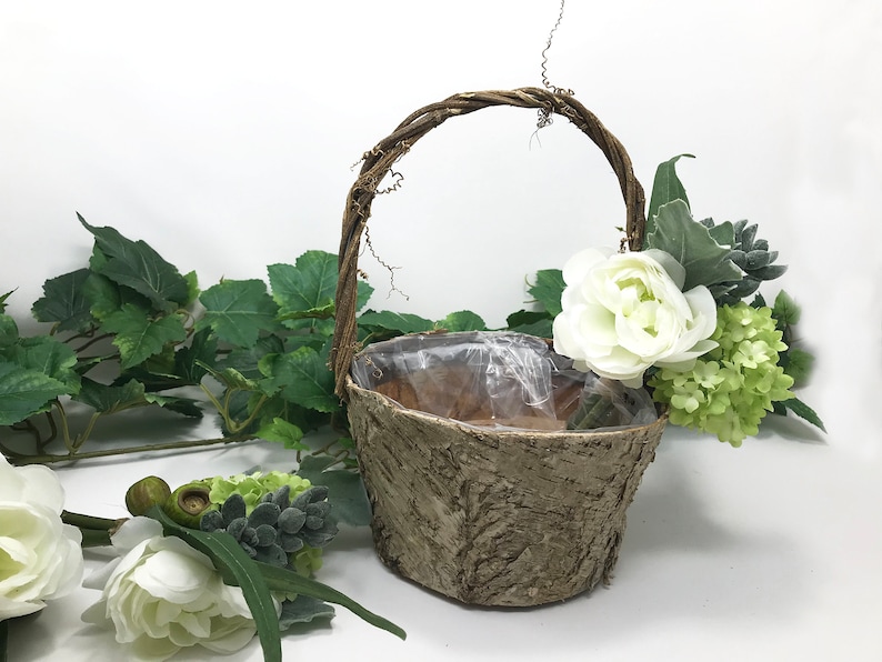 May include: A small, brown, bark-textured basket with a twig handle. The basket is filled with white flowers and green foliage.