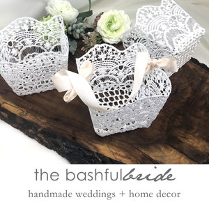 May include: Three white lace baskets with delicate floral patterns and a satin ribbon handle. The baskets are arranged on a wooden surface. The text "the bashful bride" and "handmade weddings + home decor" is visible below the baskets.