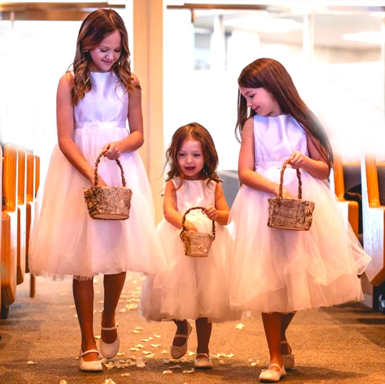 May include: Three young girls in white dresses walk down an aisle, each carrying a small woven basket. The baskets are decorated with natural elements, such as bark and twigs. The girls are smiling and looking down at their baskets.