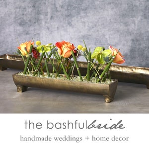 May include: A long, rectangular, gold-colored metal planter with a floral arrangement of orange roses, green leaves, and bamboo stalks. The planter has four legs and is sitting on a gray surface. The text "the bashful bride" and "handmade weddings + home decor" is below the planter.