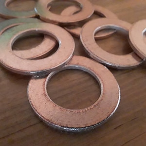 Puede incluir: Primer plano de una pila de arandelas de metal de color plateado. Las arandelas son redondas con un agujero en el centro. Están dispuestas en un patrón aleatorio sobre una superficie de madera.