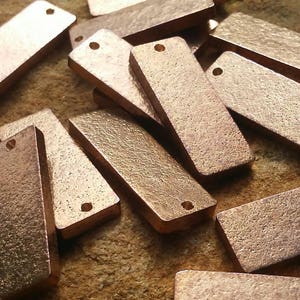 May include: A collection of rectangular copper-colored metal tags with a textured surface and a small hole at one end. The tags are scattered on a brown surface, suggesting a craft or jewelry-making theme.
