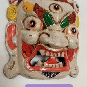 May include: A white wooden mask painted in red, yellow, green, and black. The mask has a large mouth with teeth, a third eye, and small eyes on the forehead. The mask is labelled "WMS 122A".