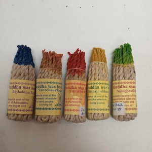 May include: Five different incense sticks with labels that say "Buddha was born" and the name of a different Buddha on each stick. The sticks are wrapped in white string and have different coloured threads woven into the string. The labels are yellow with black text.