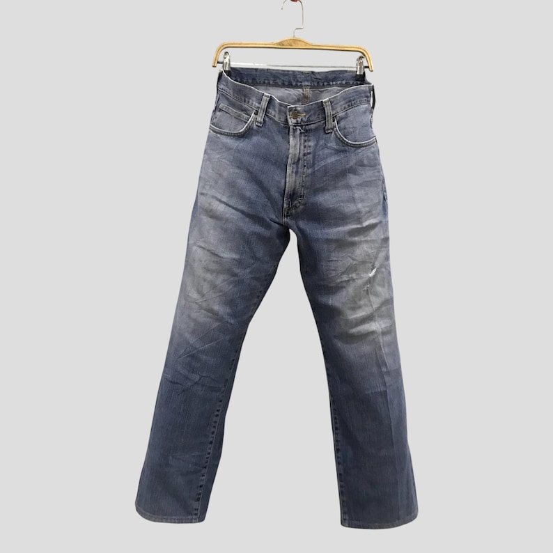 May include: Faded blue denim jeans hanging on a wooden hanger. The jeans have a classic five-pocket design, a button and zipper closure, and show signs of wear. The jeans are approximately 32 inches in length.