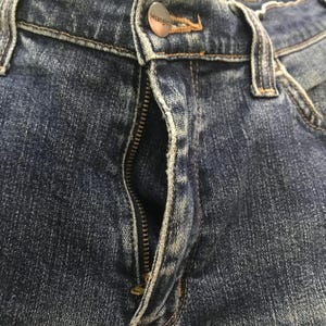 May include: Close-up of blue denim jeans with a partially unzipped fly. The jeans feature a metal button with the brand name "Wrangler" and visible stitching details. The fabric shows a worn, faded appearance.