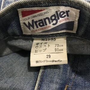 以下が含まれることがあります： 赤と白のWranglerロゴが入ったデニムジーンズのラベルのクローズアップ。ラベルには「YOUR OWN FEELING. YOUR ESSENTIAL」というテキストが含まれています。ロゴの下には、ウエスト73cm、ヒップ91cm、サイズ29の日本語のテキストと測定値が記載されたタグがあります。