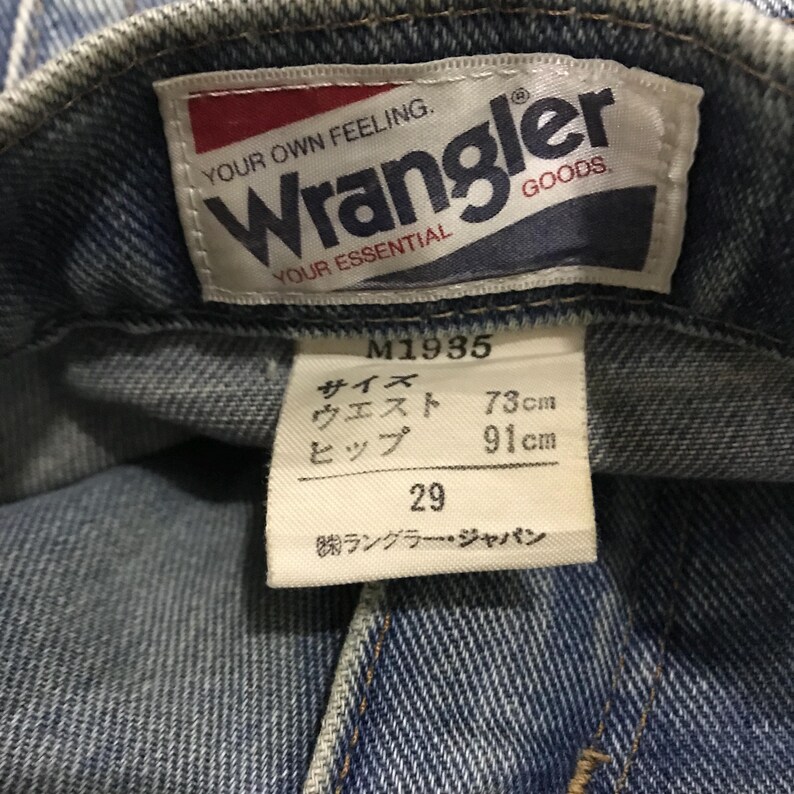 以下が含まれることがあります： 赤と白のWranglerロゴが入ったデニムジーンズのラベルのクローズアップ。ラベルには「YOUR OWN FEELING. YOUR ESSENTIAL」というテキストが含まれています。ロゴの下には、ウエスト73cm、ヒップ91cm、サイズ29の日本語のテキストと測定値が記載されたタグがあります。