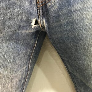 May include: Close-up of light blue denim jeans with a distressed hole near the seam. The jeans show signs of wear and tear, with visible stitching and a faded appearance. The fabric has a textured look.