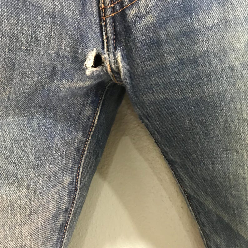 May include: Close-up of light blue denim jeans with a distressed hole near the seam. The jeans show signs of wear and tear, with visible stitching and a faded appearance. The fabric has a textured look.