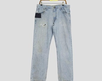 May include: Light blue denim jeans with a distressed look, featuring a patch on the left thigh and a dark blue patch near the pocket. The jeans are hanging on a wooden hanger.