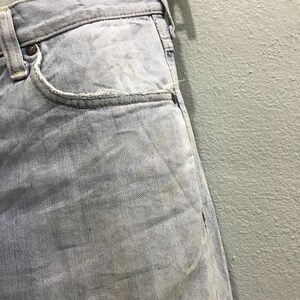 May include: Close-up of light blue denim jeans. The jeans show signs of wear and tear, with visible fading and a distressed pocket edge. The fabric has a textured appearance, with subtle variations in colour.