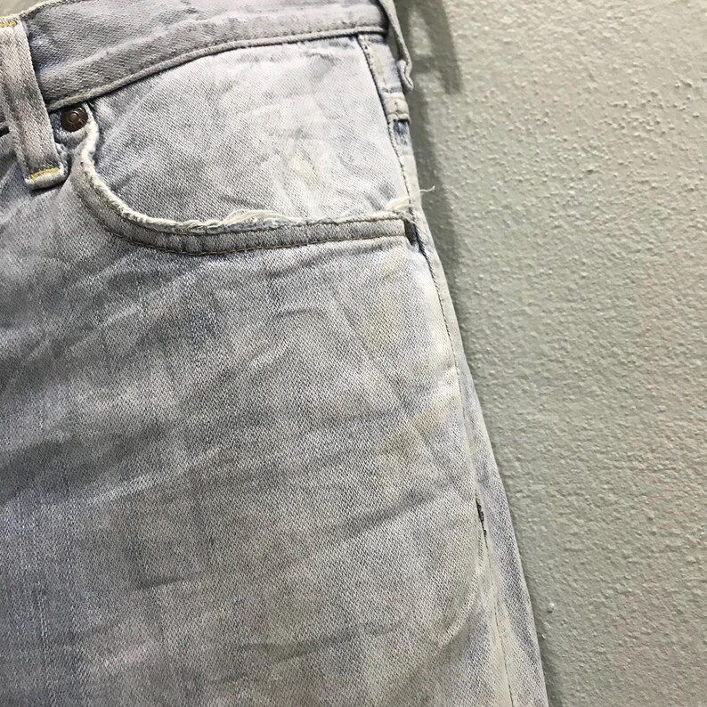 May include: Close-up of light blue denim jeans. The jeans show signs of wear and tear, with visible fading and a distressed pocket edge. The fabric has a textured appearance, with subtle variations in colour.