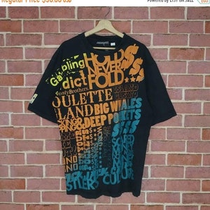 Może przedstawiać: Czarny T-shirt z krótkim rękawem z kolorowym nadrukiem graficznym z napisami "Gambling Dict Never Fold" i "Julette Island Casino Land Big Whales King & Deep Pockets".