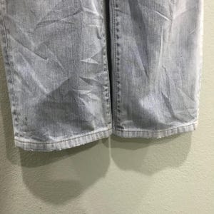 May include: Close-up of light blue denim jeans. The jeans show wear and tear, with visible creases and a faded appearance. The hem of the jeans is visible, showing the bottom edge of the fabric.
