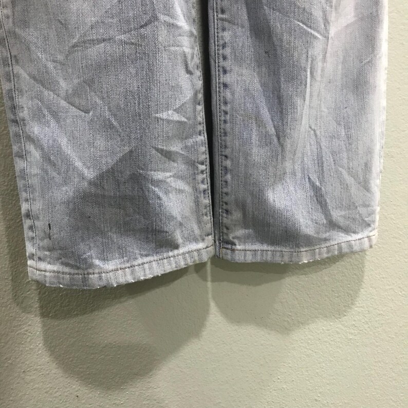 May include: Close-up of light blue denim jeans. The jeans show wear and tear, with visible creases and a faded appearance. The hem of the jeans is visible, showing the bottom edge of the fabric.