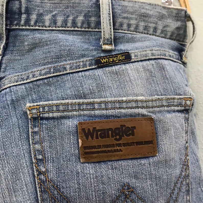 May include: Close-up of light blue denim jeans with a Wrangler label on the back pocket. The label is brown with the Wrangler logo and text. The jeans have a classic five-pocket design and a faded wash.