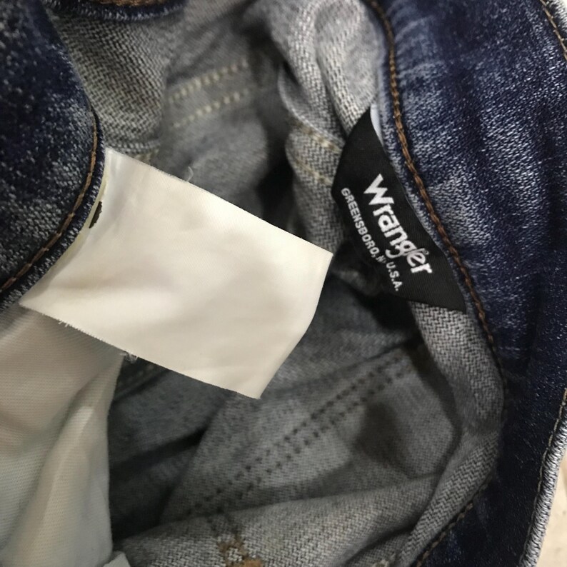 May include: Close-up of the inside of blue denim jeans. A white tag is visible, along with a black tag that reads "Wrangler Greensboro, U.S.A."