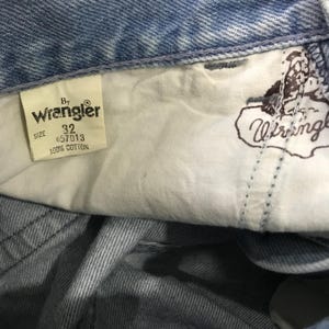 May include: Close-up of the inside of a pair of light blue denim jeans. A white tag reads "By Wrangler" with the size "32" and "100% Cotton". The Wrangler logo is also visible on the inside pocket lining.