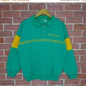 Puede incluir: Una sudadera verde de manga larga con una franja amarilla en cada manga. La sudadera tiene un cuello abotonado y el texto "AUSTRALIA" y "Hobart BRISBANE Adelaide Sydney MELBOURNE PERTH DARWIN" impreso en amarillo.