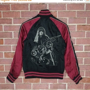 Może przedstawiać: Czarna satynowa kurtka bomber z czerwonymi rękawami i nadrukiem graficznym przedstawiającym kobietę jadącą na motocyklu. Kurtka ma biały pasek na rękawach i czarno-czerwony pas w talii.