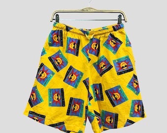 May include: Yellow shorts with a repeating pattern of a smiling sun wearing sunglasses within a blue and red square. The shorts have an elasticated waistband and are hanging on a wooden hanger.