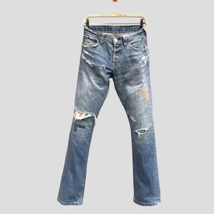 Op de afbeelding: Lichtblauwe spijkerbroek met distressed details, waaronder scheuren en patches. De jeans hangt aan een goudkleurige hanger. De jeans heeft een knoopsluiting en een klassieke vijf-zakken-stijl.
