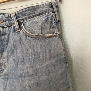 K&ouml;nnte beinhalten: Nahaufnahme einer hellblauen Jeans. Die Jeans hat eine abgenutzte Tasche mit ausgefransten Kanten. Die N&auml;hte sind in einer kontrastierenden orangefarbenen Farbe sichtbar. Die Jeans h&auml;ngt an einem Metallb&uuml;gel.