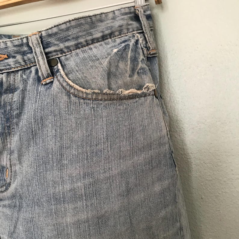 K&ouml;nnte beinhalten: Nahaufnahme einer hellblauen Jeans. Die Jeans hat eine abgenutzte Tasche mit ausgefransten Kanten. Die N&auml;hte sind in einer kontrastierenden orangefarbenen Farbe sichtbar. Die Jeans h&auml;ngt an einem Metallb&uuml;gel.