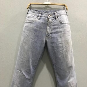 May include: Light blue denim jeans hanging on a wooden hanger. The jeans have a classic five-pocket design, a zip fly, and a faded wash. The jeans are a light blue colour.