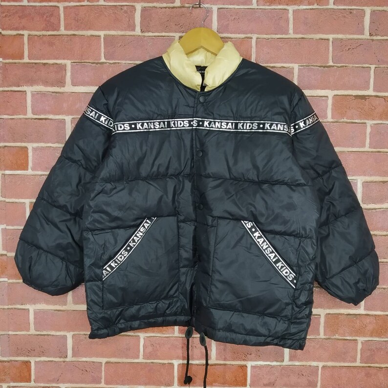 May include: Black puffer jacket with a white zipper and a white band around the collar. The band has the text "KANSAI KIDS" repeated on it.
