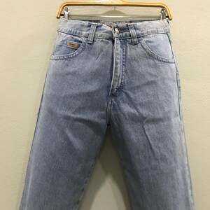 May include: Light blue denim jeans hanging on a wooden hanger. The jeans have a classic five-pocket design, a button and zipper closure, and a small brown label on the front pocket. The jeans are made of a light-wash denim fabric.