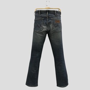 Pu&ograve; includere: Jeans in denim blu scuro appesi a una gruccia dorata. I jeans hanno un classico design a cinque tasche, una toppa in pelle marrone e una gamba dritta. I jeans sono di colore blu sbiadito.