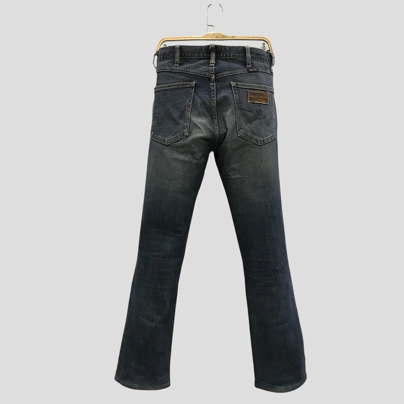 Pu&ograve; includere: Jeans in denim blu scuro appesi a una gruccia dorata. I jeans hanno un classico design a cinque tasche, una toppa in pelle marrone e una gamba dritta. I jeans sono di colore blu sbiadito.