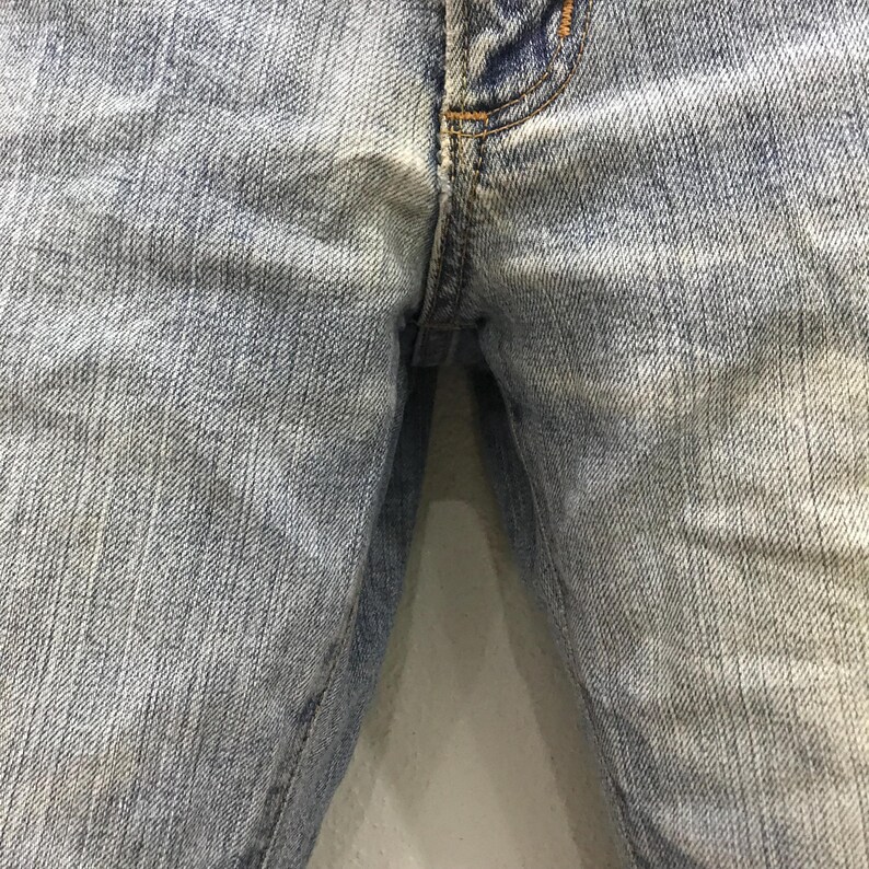 May include: Close-up of light blue denim jeans. The jeans show wear and tear, with visible fading and subtle discoloration. The image focuses on the front of the jeans, highlighting the button and zipper area.