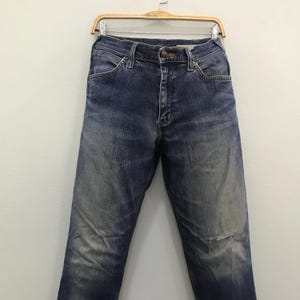 May include: Blue denim jeans with a faded wash and a distressed knee. The jeans are hanging on a wooden hanger against a white background. The jeans have a classic five-pocket design.