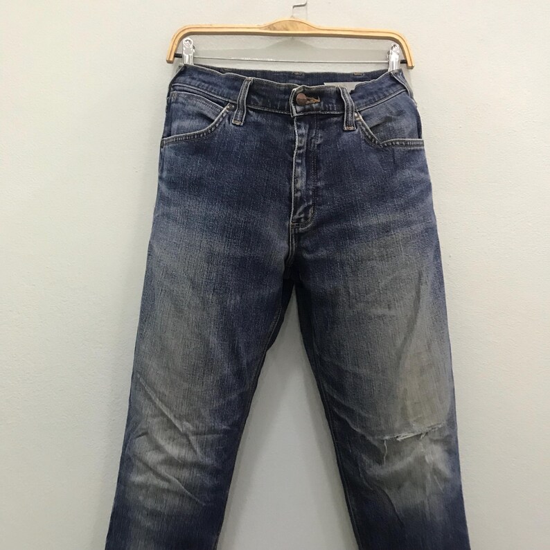 May include: Blue denim jeans with a faded wash and a distressed knee. The jeans are hanging on a wooden hanger against a white background. The jeans have a classic five-pocket design.