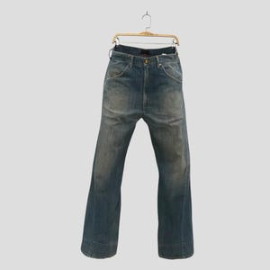 Peut inclure: Un jean en denim bleu avec un délavage usé, une taille haute et des jambes larges. Le pantalon comprend une braguette à boutons, cinq poches et des passants de ceinture, présenté suspendu à un cintre en bois sur un fond uni.