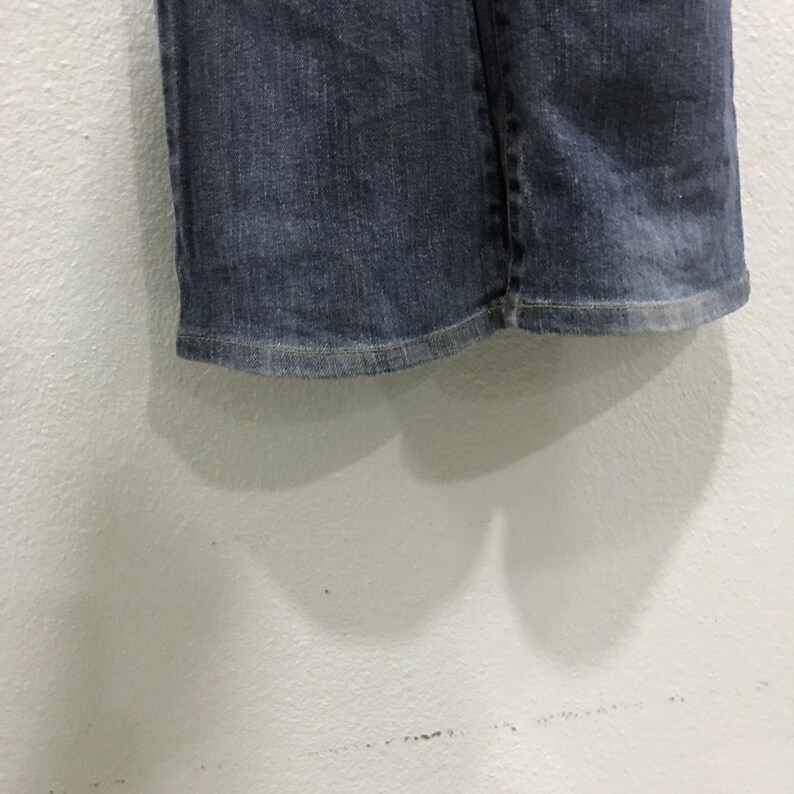 Pu&ograve; includere: Primo piano dell'orlo di un paio di jeans blu. I jeans sono di un lavaggio medio con un aspetto leggermente sbiadito. L'orlo &egrave; cucito con cura con un filo chiaro e il tessuto mostra una sottile usura.