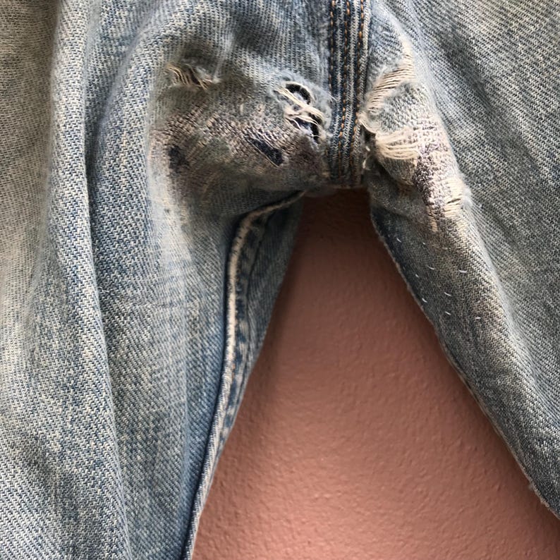 May include: Close-up of light blue denim jeans with distressed details. The jeans show visible wear and tear, with multiple rips and frayed edges around the crotch area. The fabric has a faded appearance, suggesting a vintage or well-worn style.