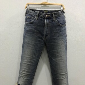 Pu&ograve; includere: Jeans in denim blu scuro con un classico design a cinque tasche. I jeans sono appesi a una gruccia di legno su uno sfondo bianco. I jeans hanno una chiusura con bottoni e un aspetto sbiadito e usurato.